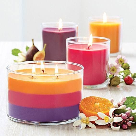 Lit colorful candles in glass jars paired with fresh orange slices, figs, and white blossoms.